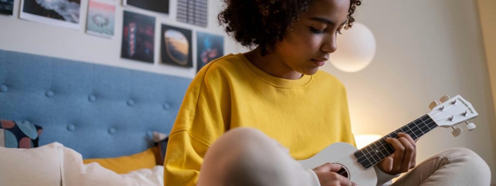 A young woman in a yellow sweatshirt playing a white ukulele on her bed, focusing on learning the instrument as she chooses the best musical instrument for her style.