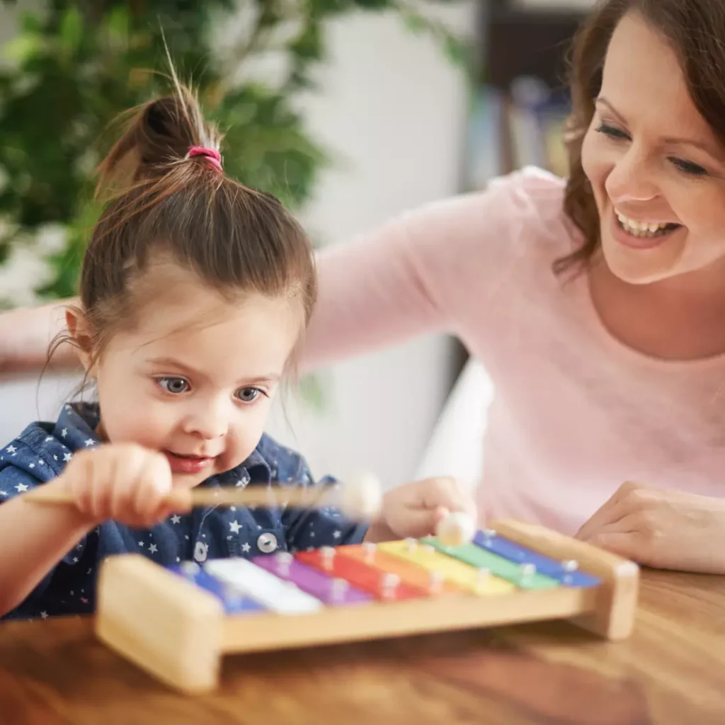 Cute girl playing Goldesi’s rainbow xylophone on a table, with her mother smiling in the background